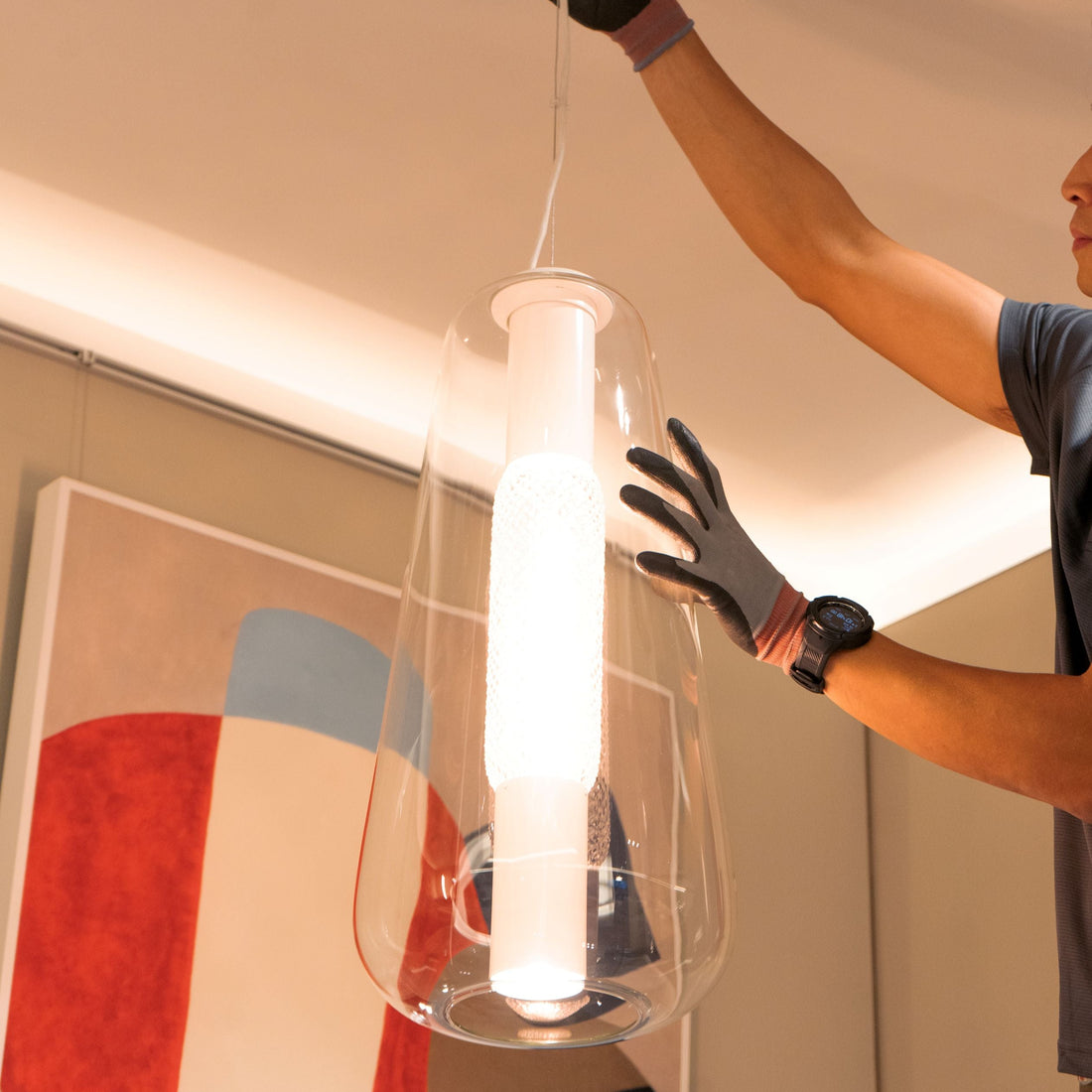 Person installing a modern pendant light fixture in a room with abstract art on the wall.